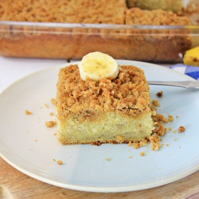 A square slice of crumb-topped banana coffee cake sits on a white plate, garnished with a banana slice; the rest of the cake is in a glass baking dish in the background.