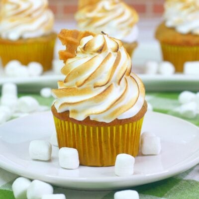 A cupcake with toasted meringue frosting sits on a white plate surrounded by mini marshmallows, with more cupcakes in the background.