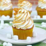 A cupcake with toasted marshmallow frosting in a yellow wrapper sits on a plate surrounded by small marshmallows.