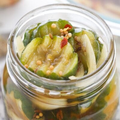 A close-up of sliced pickles with seeds and spices in a glass jar filled with brine.