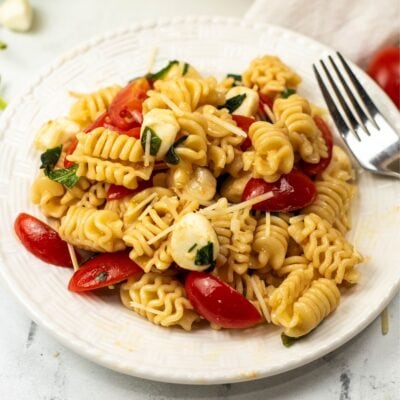 A white plate with pasta salad containing radiatore pasta, cherry tomatoes, mozzarella balls, fresh basil, and grated parmesan cheese, with a fork on the plate.