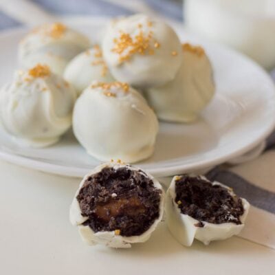 A plate of white chocolate-coated truffles with a cut-open truffle in front, showing a dark chocolate filling and caramel center, topped with gold sprinkles.