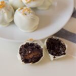 A plate of white chocolate-coated truffles, with one truffle cut open to reveal a dark interior and a caramel center, topped with gold sprinkles.