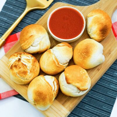 A wooden board with eight baked bread rolls and a bowl of red dipping sauce, placed on a blue-striped surface with a gold serving utensil nearby.