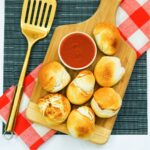 A wooden board with seven baked rolls and a bowl of red dipping sauce, next to a gold spatula and a red-and-white checkered napkin.