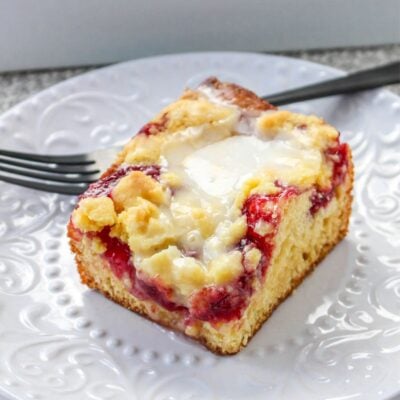 A slice of crumb-topped coffee cake with a layer of fruit filling and cream cheese on a white decorative plate, with a black fork in the background.