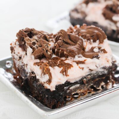 A close-up of a chocolate brownie topped with a layer of pink cream and drizzled chocolate, served on a silver square plate.