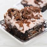 A square piece of chocolate cake topped with pink frosting and drizzled with chocolate on a silver plate.