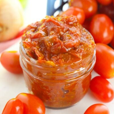 A small glass jar filled with chunky tomato sauce is surrounded by fresh grape tomatoes on a white surface.