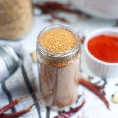A glass jar filled with a reddish-brown spice blend sits on a striped cloth, with dried chili peppers and a bowl of ground red spice nearby.