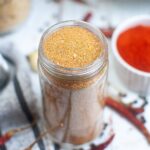 A glass jar filled with a reddish-brown spice blend sits on a table, with a small bowl of red powder in the background.