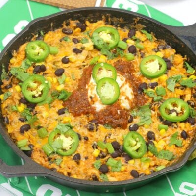 A skillet filled with a cheesy rice dish topped with black beans, corn, sliced jalapeños, salsa, sour cream, green onions, and cilantro.