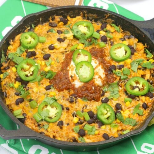 A skillet filled with a cheesy rice dish topped with black beans, corn, sliced jalapeños, salsa, sour cream, green onions, and cilantro.