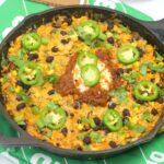 A skillet filled with cheesy rice, black beans, corn, and vegetables, topped with sour cream, salsa, sliced jalapeños, and cilantro, on a green football-themed tablecloth.