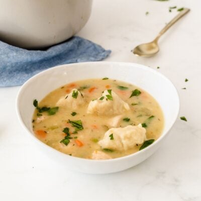A white bowl of creamy chicken and dumplings soup with chopped parsley on top, placed on a white surface next to a spoon and a blue napkin.