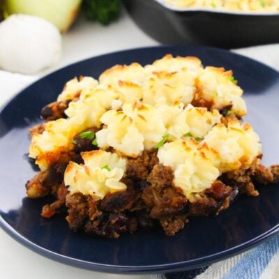A serving of shepherd's pie with browned mashed potatoes on top of ground meat and vegetables, presented on a dark plate.