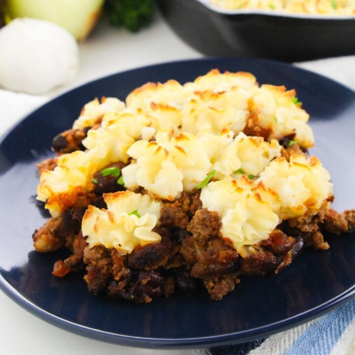 A serving of shepherd's pie with browned mashed potatoes on top of ground meat and vegetables, presented on a dark plate.