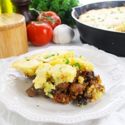 A serving of cornbread casserole with ground meat and vegetables on a white plate, garnished with parsley; fresh tomatoes and a skillet are in the background.