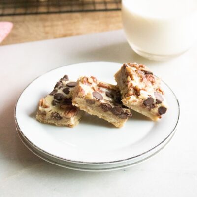 Three dessert bars with chocolate chips and nuts on a white plate, with a glass of milk in the background.