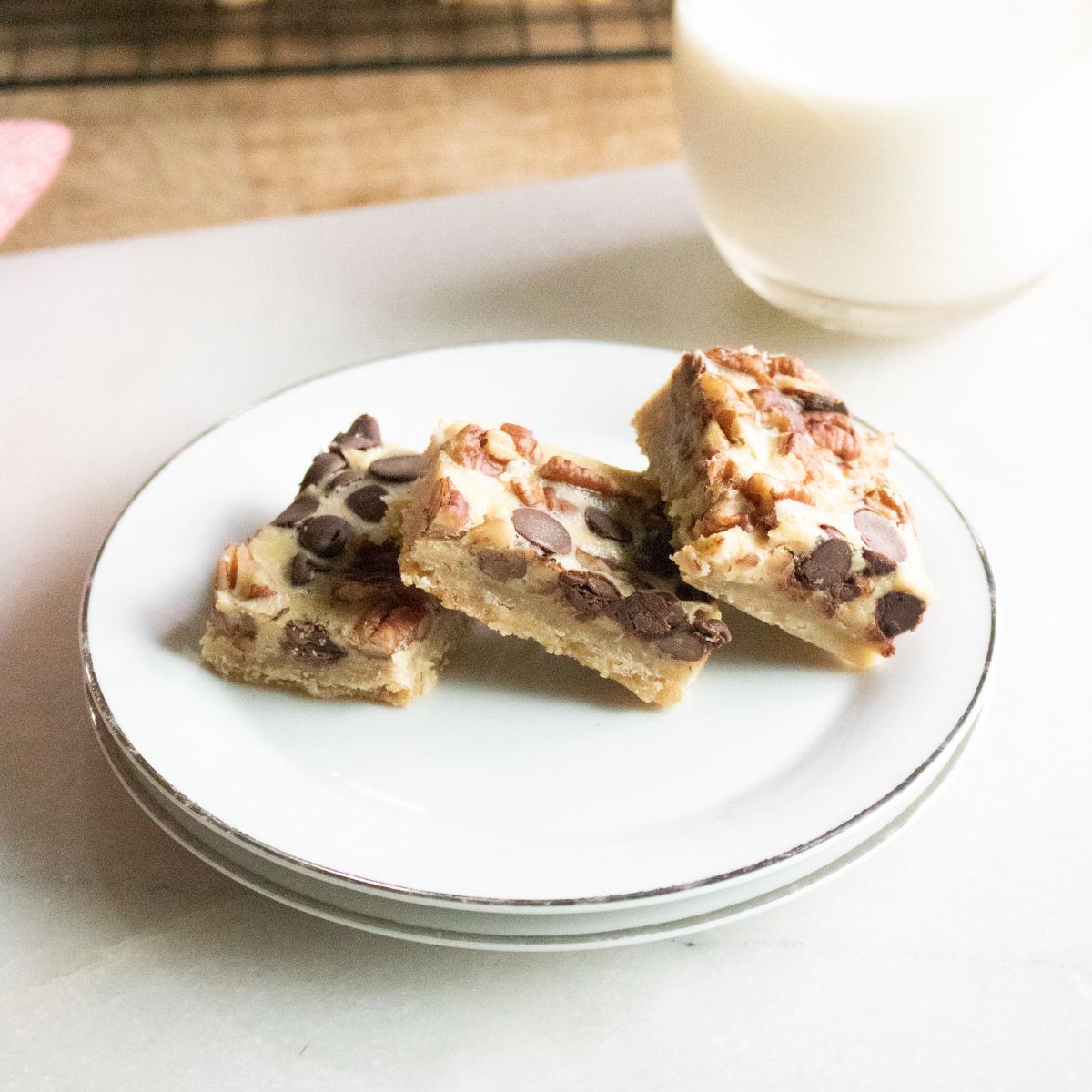 Three dessert bars with chocolate chips and nuts on a white plate, with a glass of milk in the background.