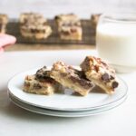 Three dessert bars with chocolate chips and nuts on a small plate, with a glass of milk and more bars blurred in the background.