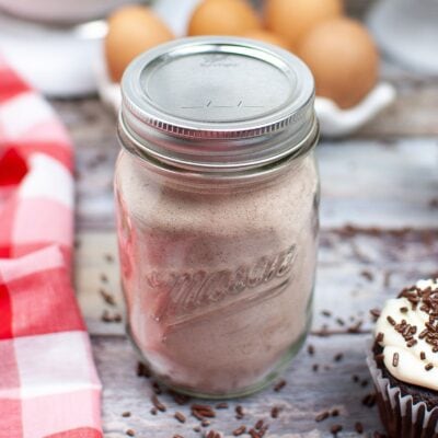 A glass mason jar filled with light brown powder sits on a wooden table next to a red and white checkered cloth, eggs, and a decorated cupcake.