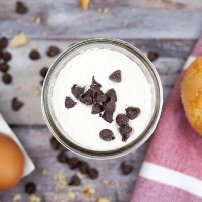 A glass jar filled with cream topped with chocolate chips, surrounded by chocolate chips, an egg, and a muffin on a wooden surface.