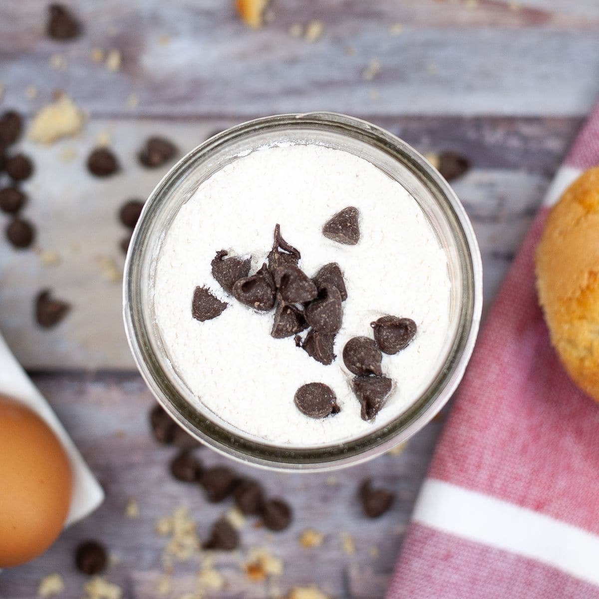 A glass jar filled with cream topped with chocolate chips, surrounded by chocolate chips, an egg, and a muffin on a wooden surface.