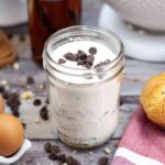 A mason jar filled with a white powdered mixture and chocolate chips on top, surrounded by eggs, chocolate chips, and a muffin on a wooden surface.