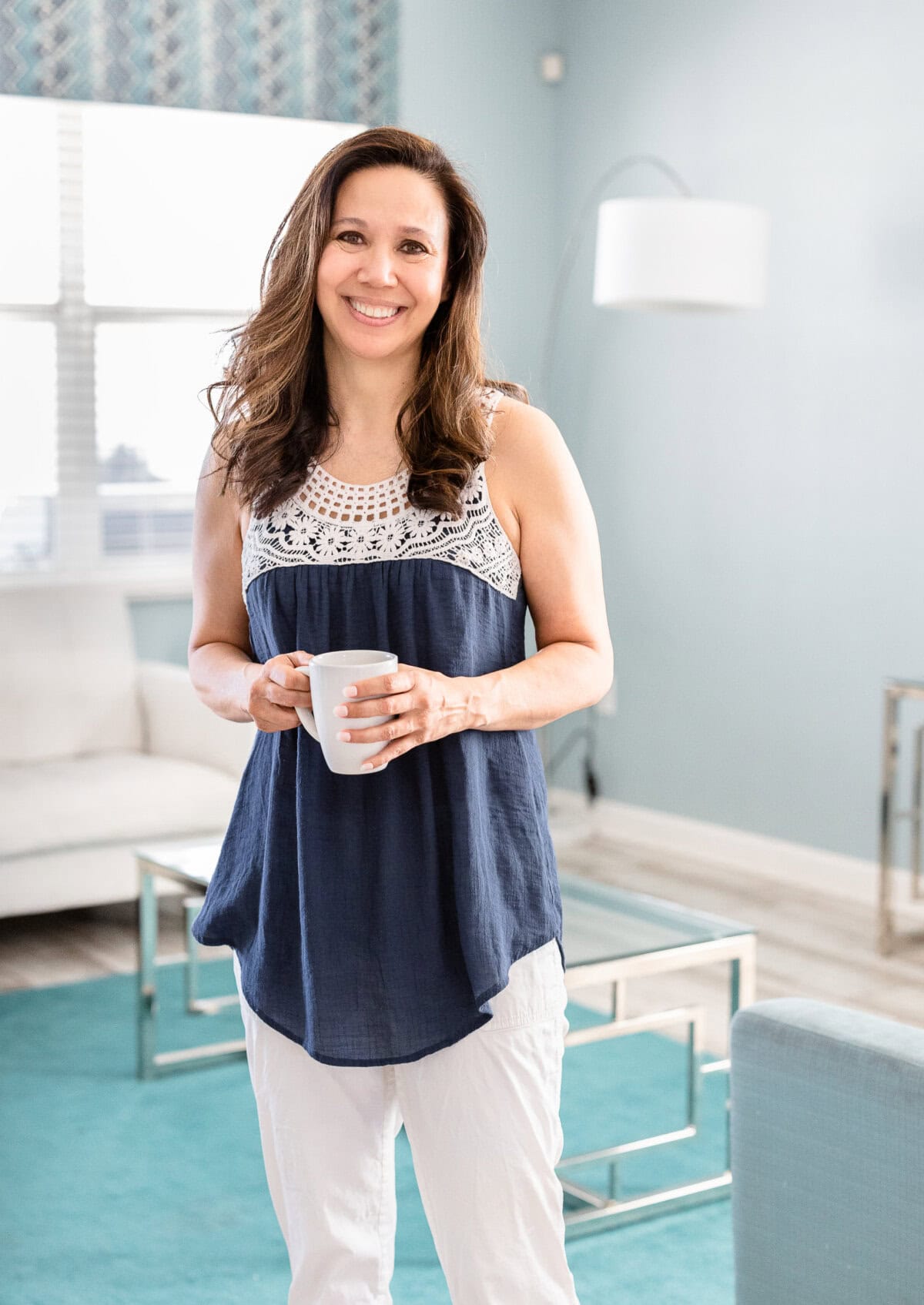 A woman stands in a bright living room holding a white mug, wearing a navy sleeveless top with white lace and white pants, smiling at the camera.