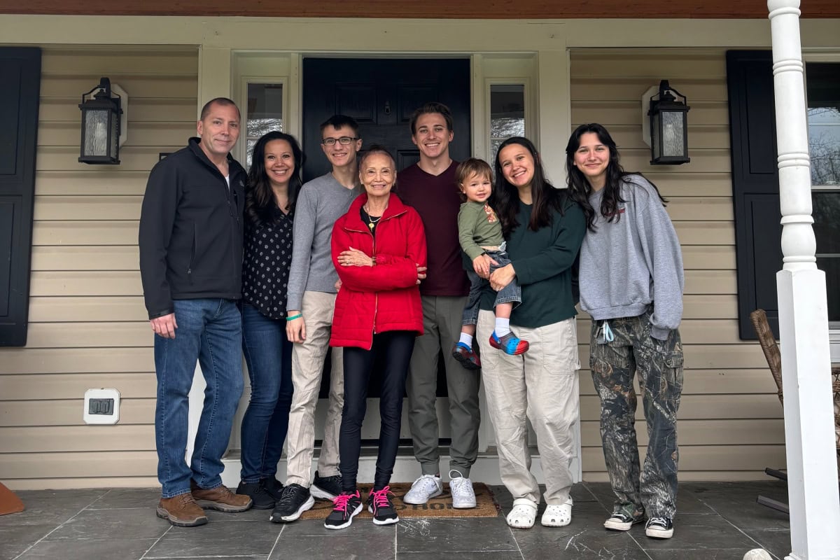 Seven adults and one toddler stand together and smile on the front porch of a house with beige siding and black shutters.